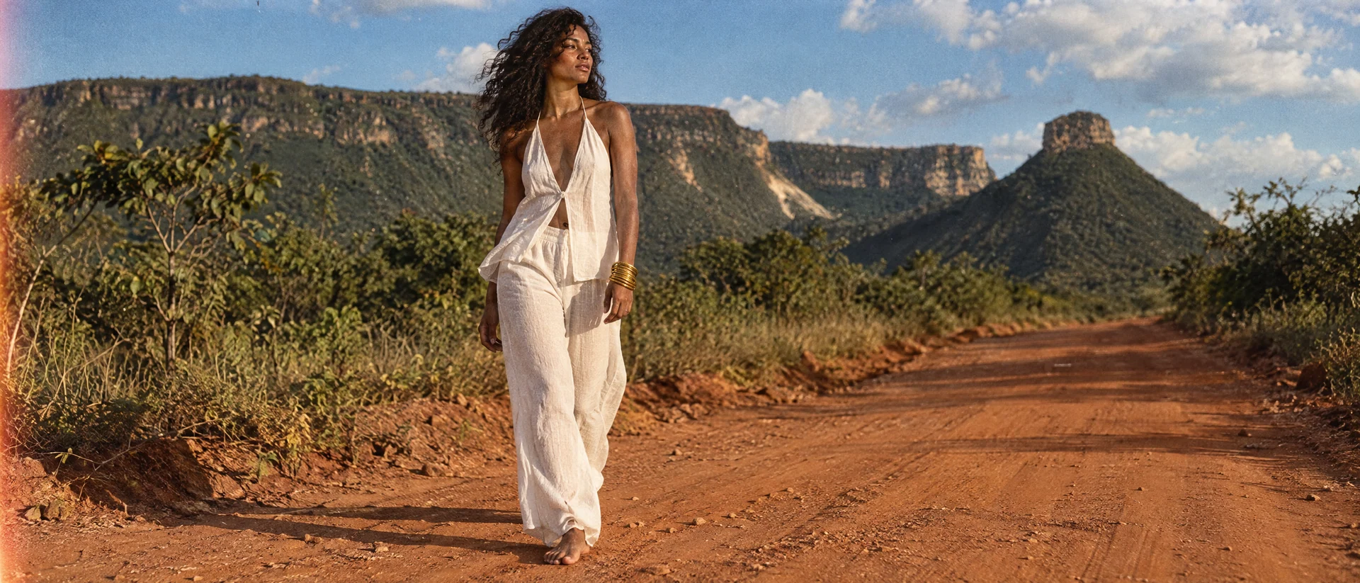 A model in a pale outfit on a sunlit path in a rocky landscape.